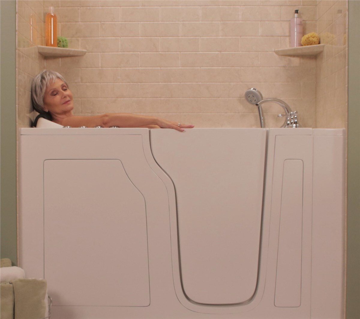 A person reclining in a white walk-in bathtub with beige tiled walls and shelves holding bath products.