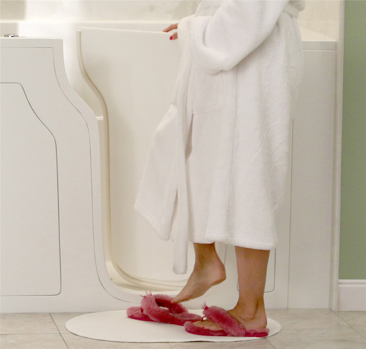 A person in a white bathrobe standing near the open door of a walk-in bathtub, wearing pink slippers on a white mat.