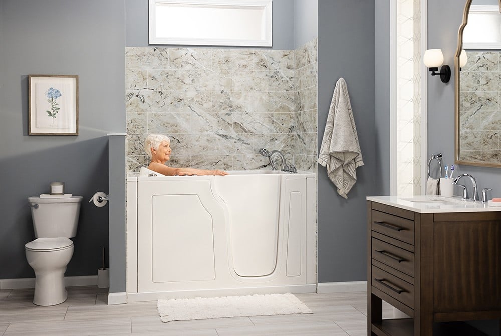 A modern bathroom with a walk-in bathtub featuring a white door and chrome fixtures, set against a marble-patterned tile wall next to a vanity and toilet.
