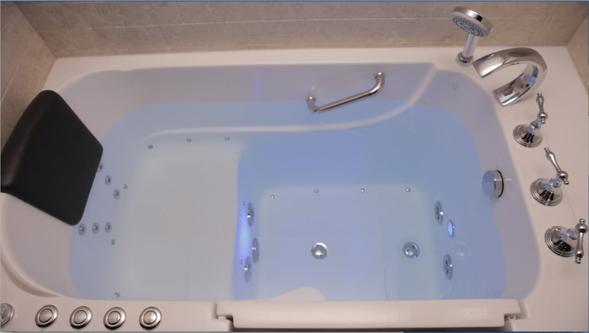 Overhead view of a walk-in bathtub partially filled with clear water, featuring a black headrest, chrome fixtures, and multiple jets.