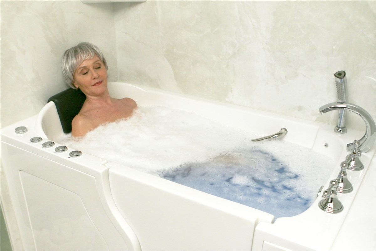 A walk-in bathtub filled with foamy water, featuring chrome fixtures and a black headrest against a light-colored wall.