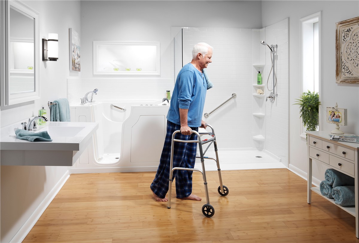 A modern bathroom with a walk-in bathtub and a separate shower area, featuring white subway tiles and wooden flooring. A person is using a walker near the bathtub, and the room includes a vanity, sink, and decorative shelving.