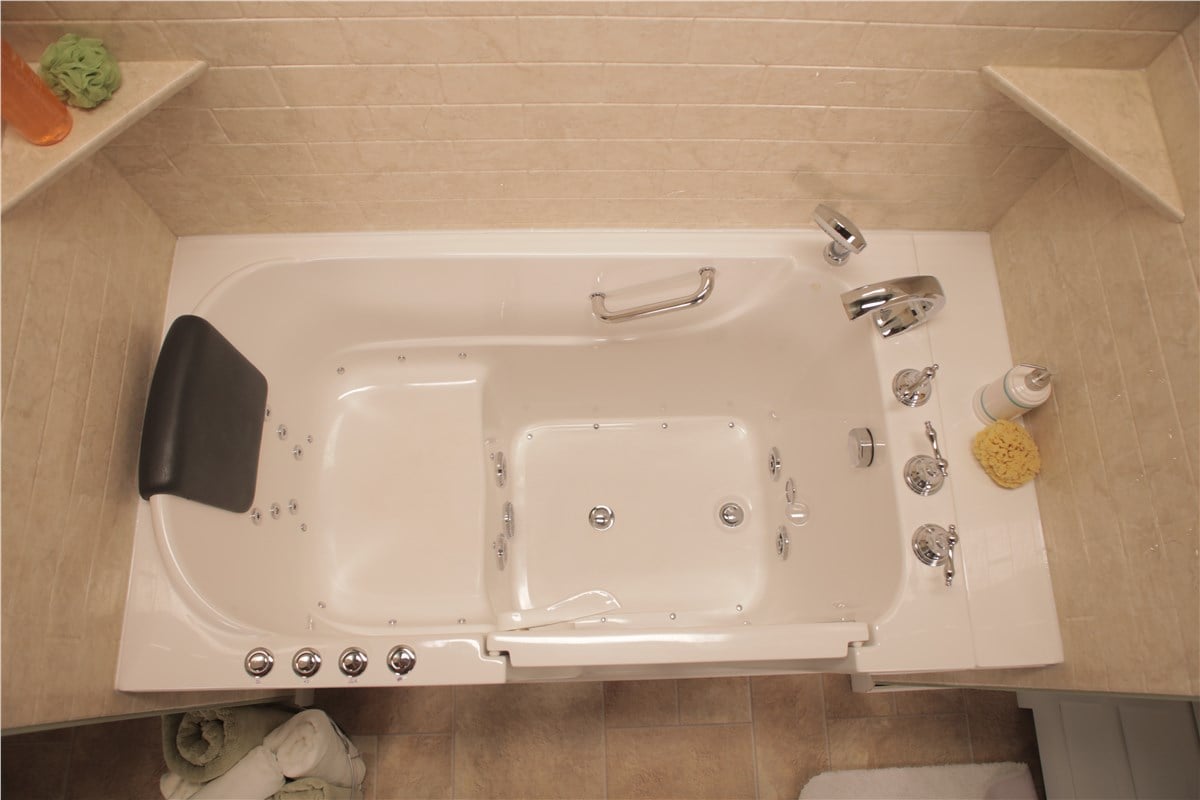 Overhead view of a white walk-in bathtub with a black headrest, chrome fixtures, and multiple jets, installed against beige tiled walls.