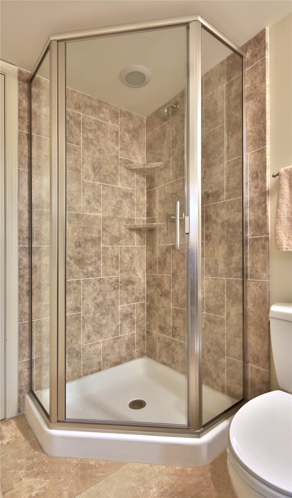 A compact bathroom with a corner glass shower, beige marbled tiles, and adjacent toilet with towel rack.