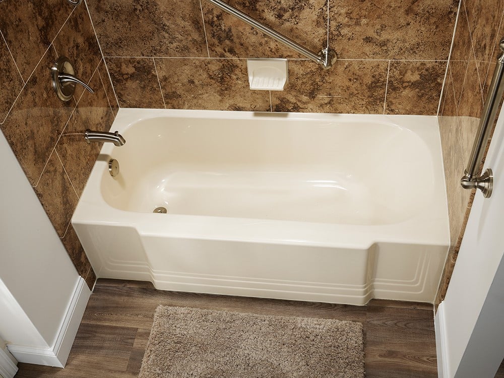 A practical bathroom with a beige bathtub, marbled tiled walls, diagonal grab bar, and wood-look flooring.